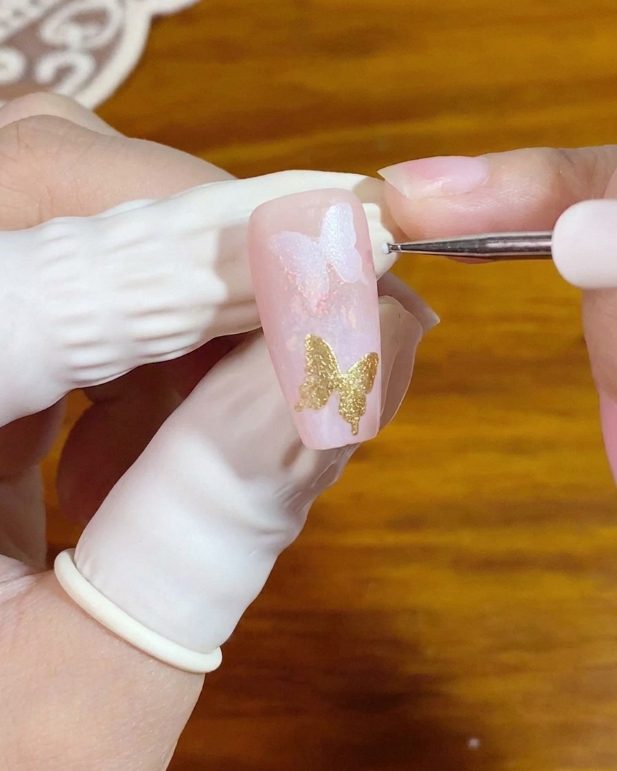 A fine detail brush applying tiny white dots next to a gold butterfly nail art element.