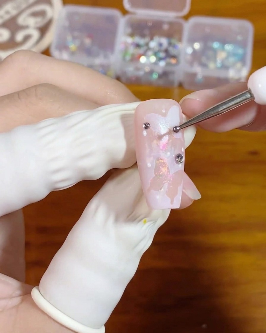 A close-up of a hand using a fine dotting tool to place a tiny bead of clear gel onto a pink nail decorated with white butterfly art.