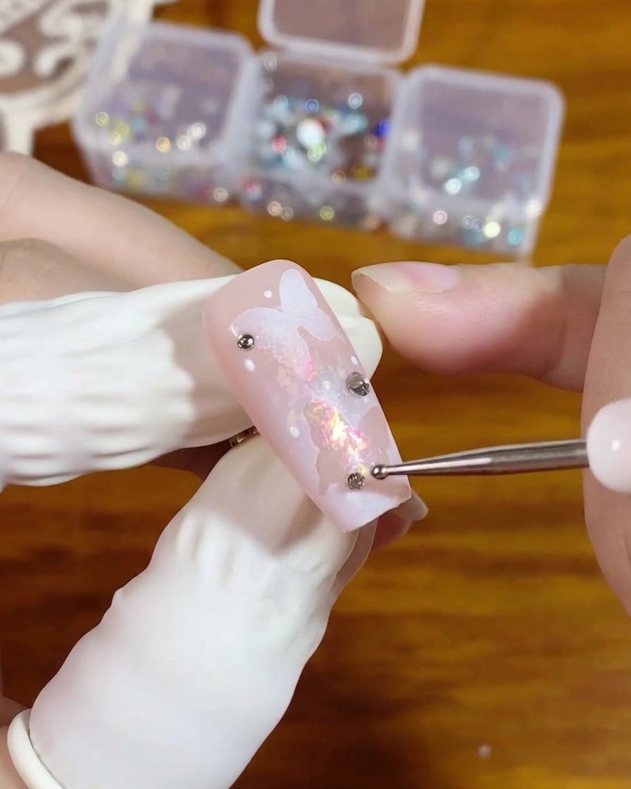 A dotting tool placing a small flat-back silver rhinestone onto a bead of clear gel on a pink nail featuring a butterfly design.