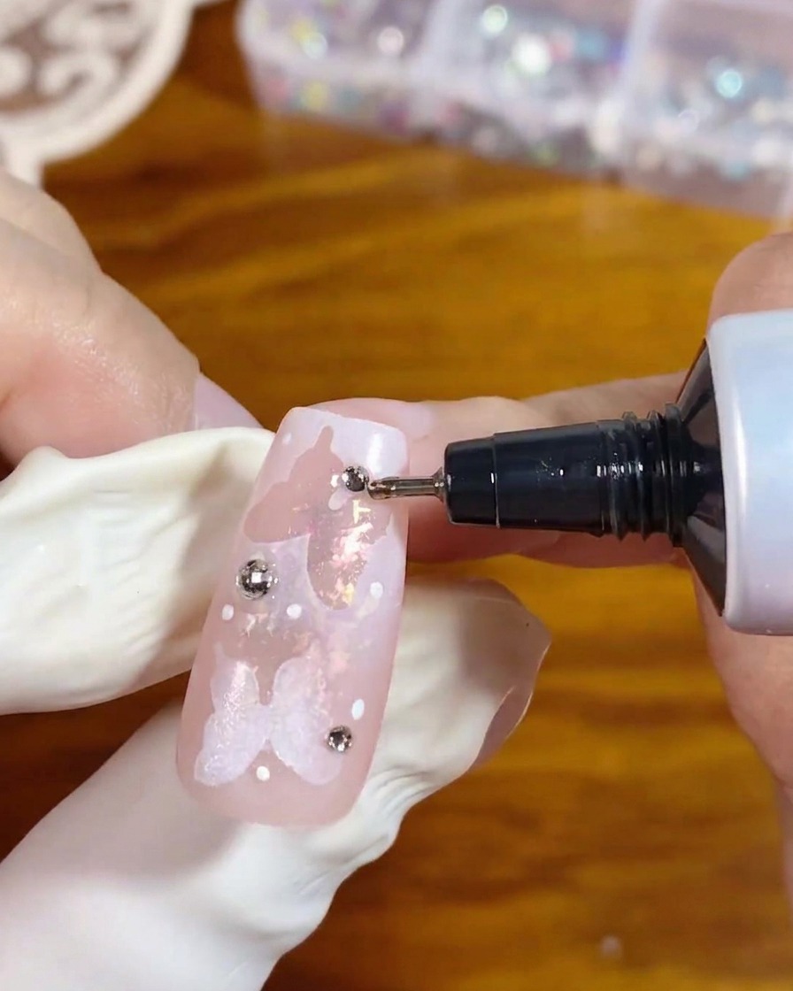 A precision tip applicator tube dispensing clear gel around the base of a silver rhinestone on a decorated pink nail to secure it in place.
