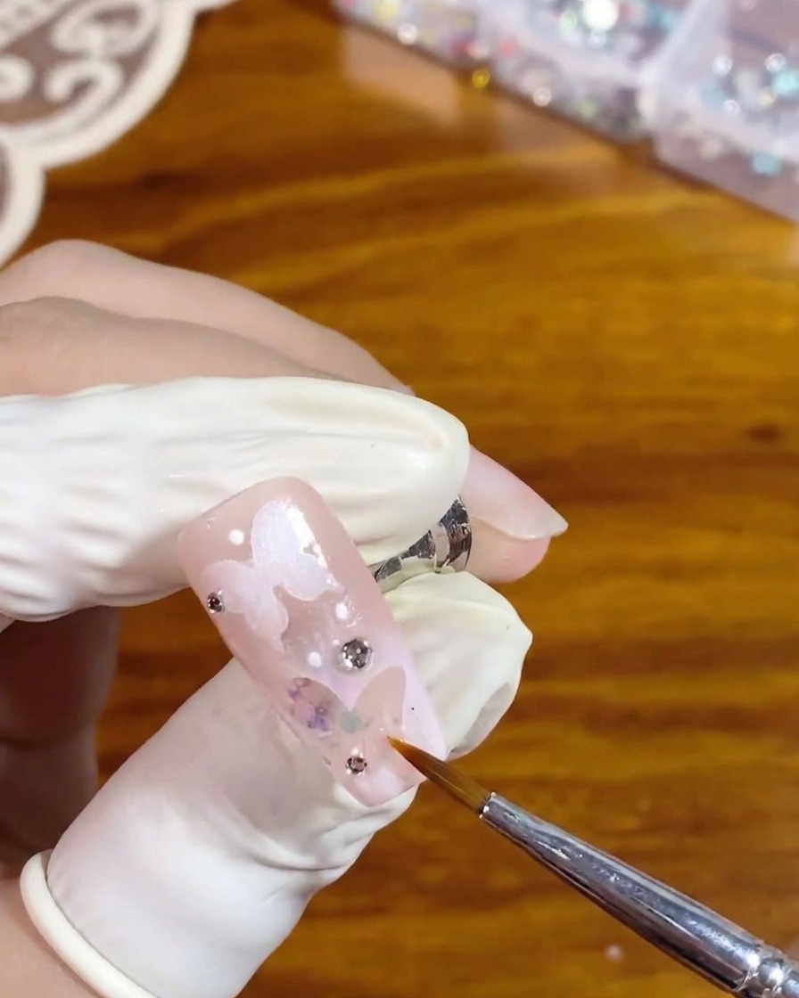 A fine liner brush carefully applying clear glossy top coat around a silver rhinestone on a pink nail with a white hollow butterfly design.