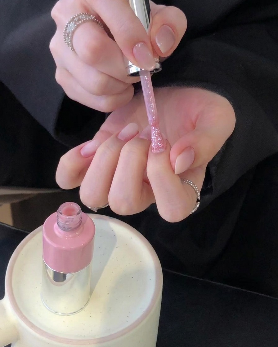 A pair of hands holding a pink polish brush over a set of manicured nails with a pink bottle resting on a ceramic holder.