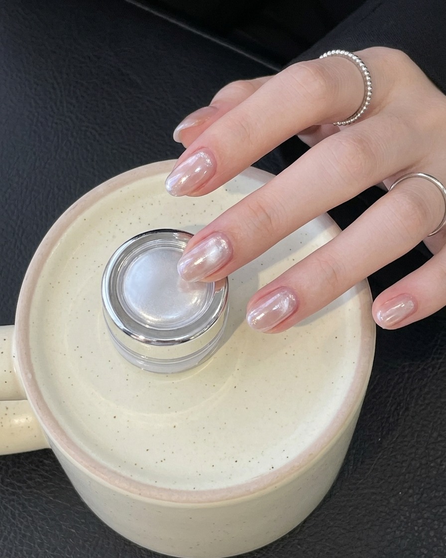 A hand wearing a ring holding a small silver jar while showcasing finished glossy chrome nails.