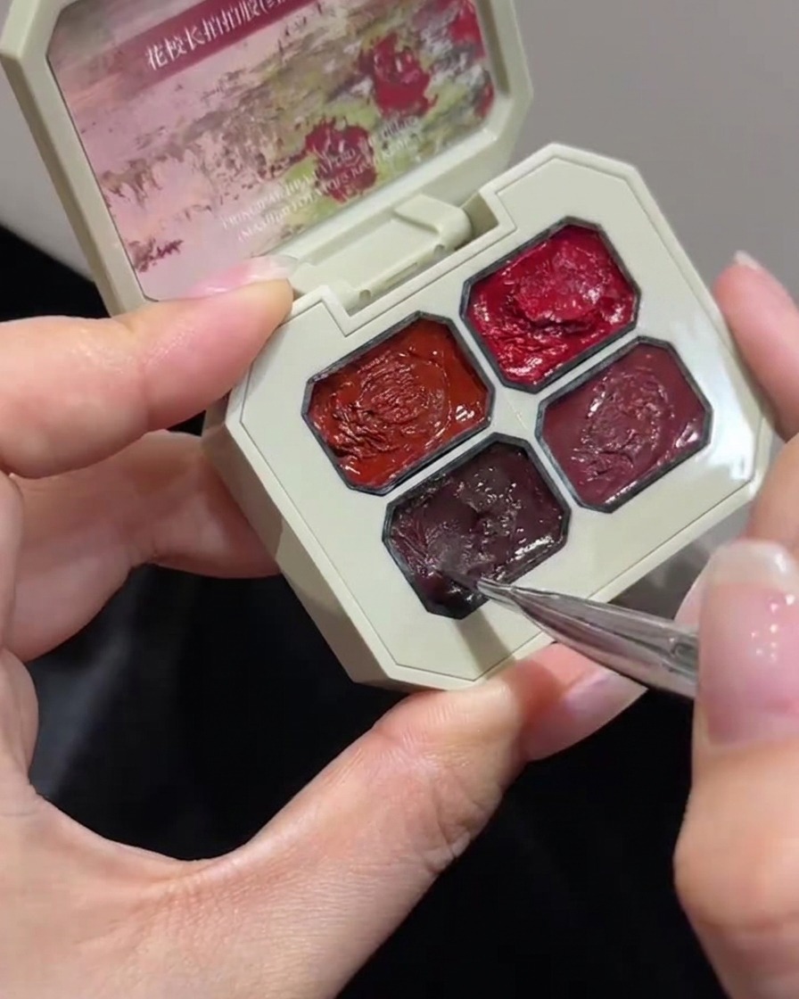 A close-up shot of a fine nail art brush dipping into a specific compartment of a multi-well rose colored gel palette.