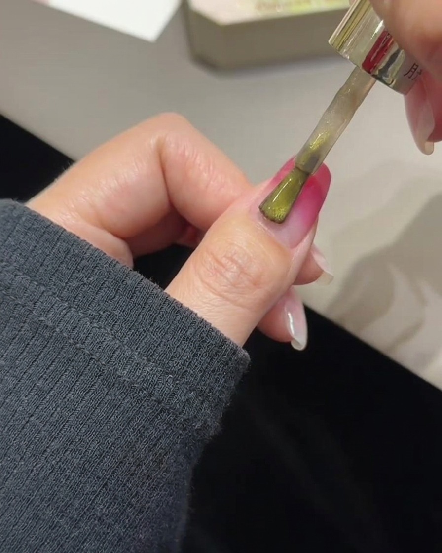 A brush applying golden cat eye gel over a cured rose gradient nail.