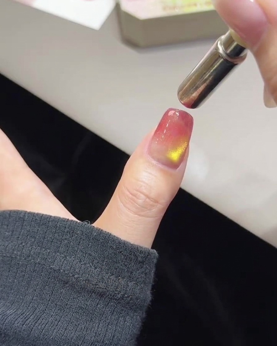 A close-up of a thumb with uncured pink and gold polish and a cylindrical magnetic wand held hovering at the nail tip.