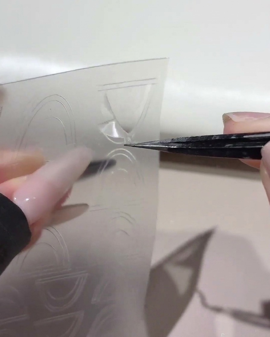 A pair of tweezers lifting a curved French guide sticker from a clear plastic sheet.