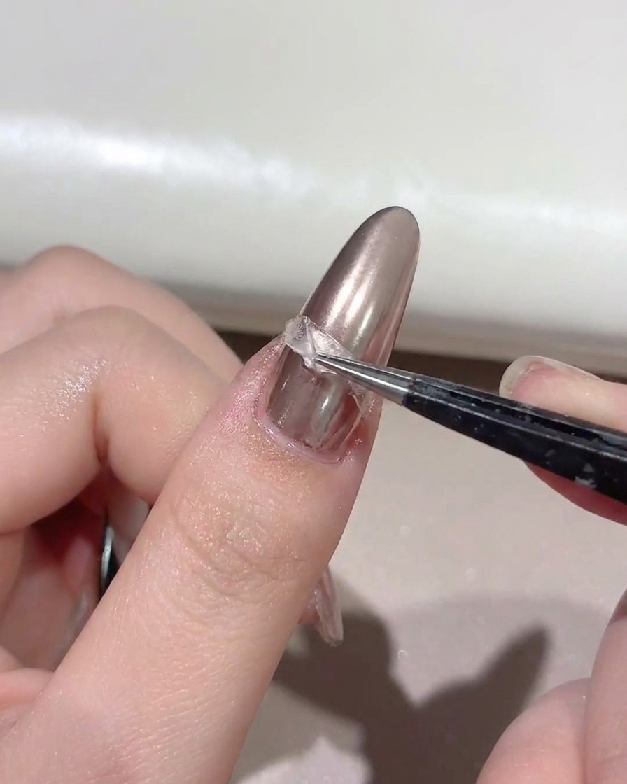 A pair of tweezers lifting a curved guide sticker off a nail to reveal a metallic line.