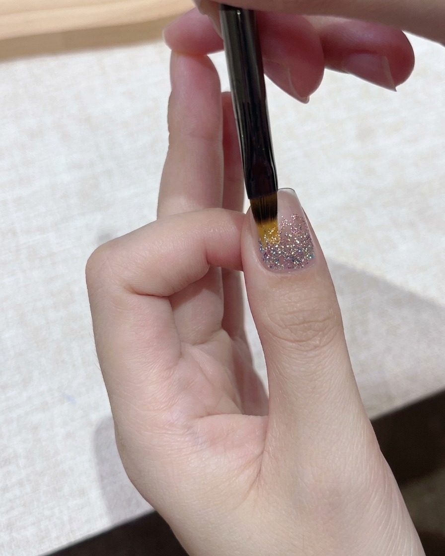 A close-up view of a fine detail brush applying a clear gel layer over a sparkling glitter nail foundation.