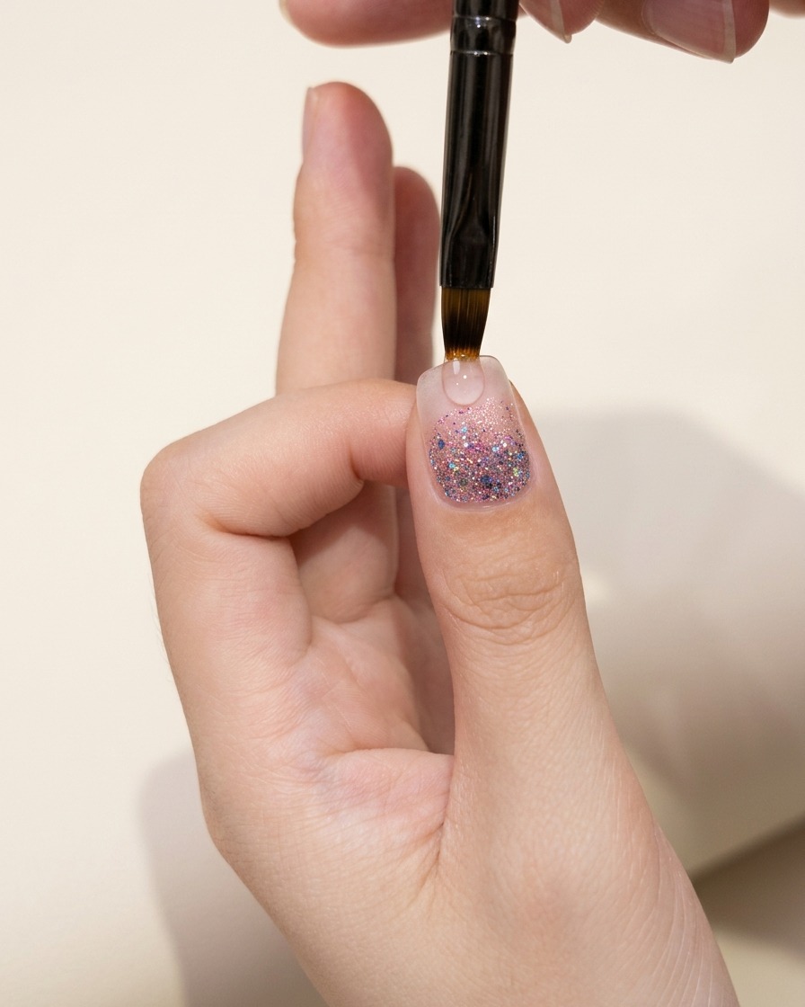 A close-up of a thumb nail showing a glitter gradient layer inspected under warm lighting.
