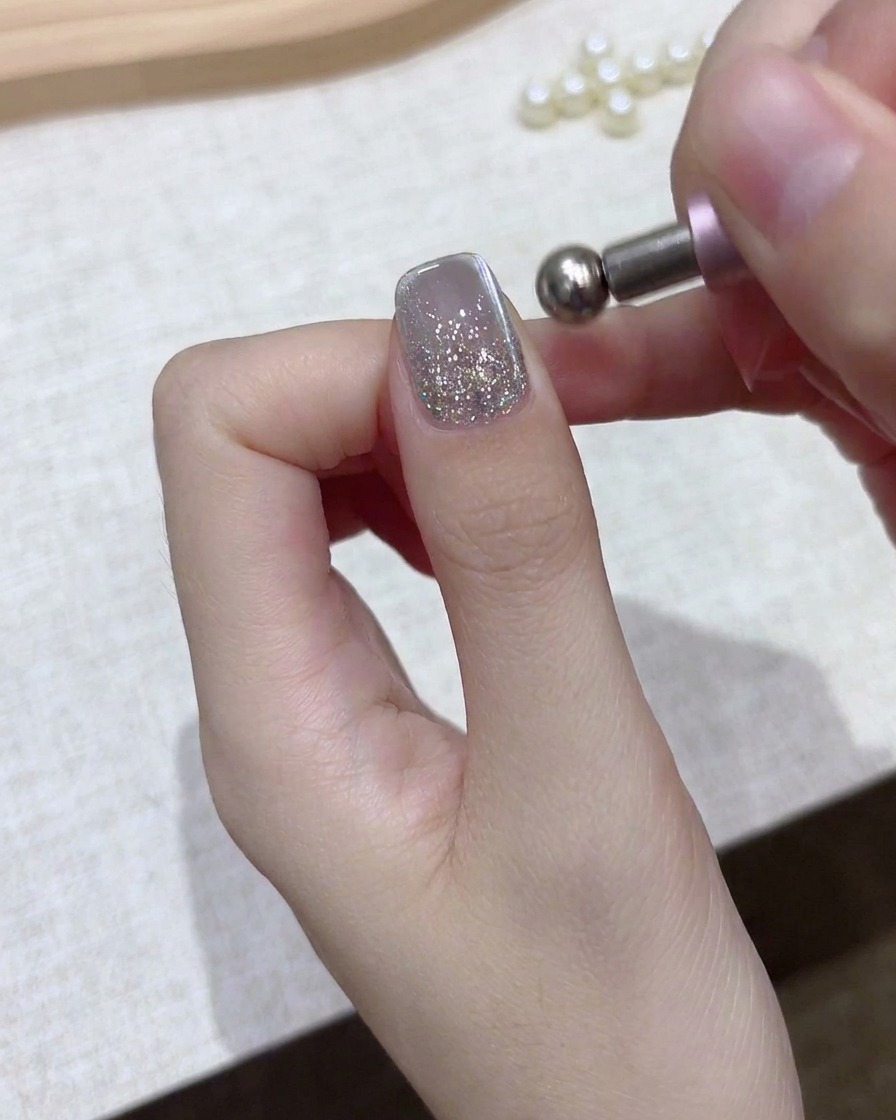 A close-up of a hand holding a spherical magnetic tool over a silver glitter gel nail.