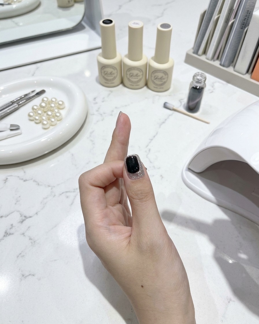 A nail brush applying a glossy top coat over a finished cat-eye nail with manicure tools and polish bottles on a white marble table in the background.