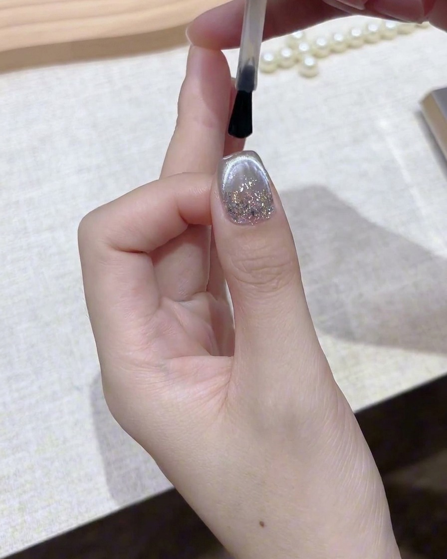 A brush applying a clear, glossy top coat over a finished silver magnetic gel nail.