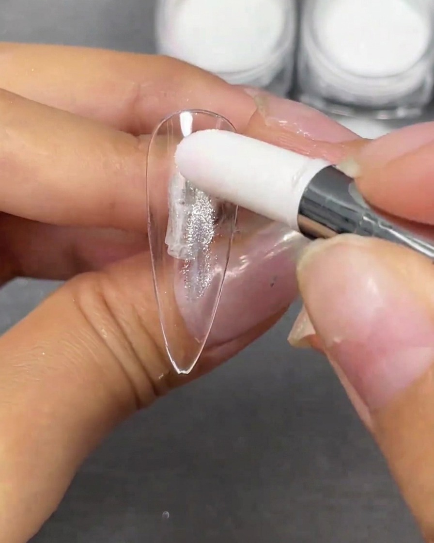 A hand using a white sponge applicator to dab silver crystal plating powder onto a clear stiletto nail tip.