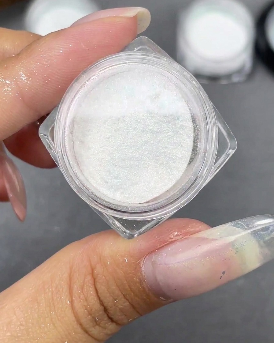 A small jar of white Moonlight powder open next to a clear stiletto nail tip.