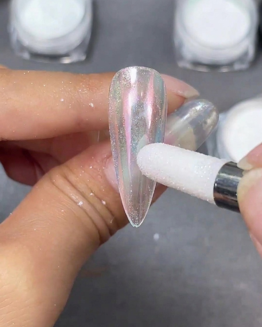 A white sponge applicator burnishing pearlescent moonlight powder onto a clear stiletto nail tip, creating an iridescent shift.