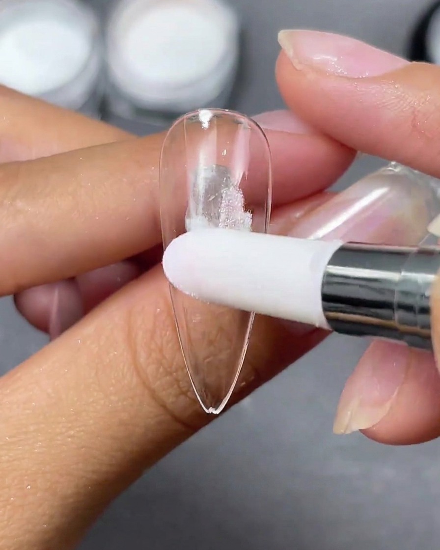 A sponge applicator dabbing white Aurora powder onto a clear stiletto nail tip.