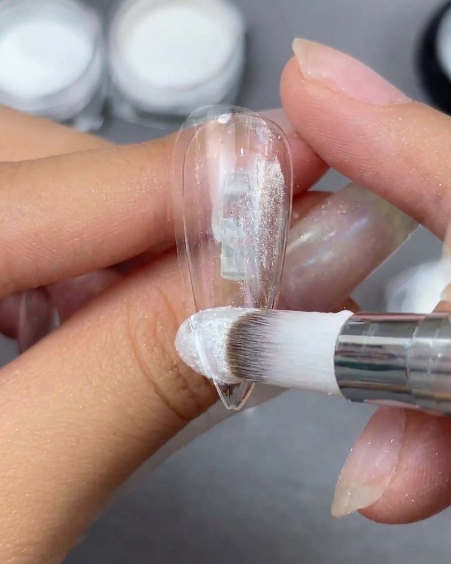A sponge applicator buffing Aurora powder onto a clear nail tip to reveal an iridescent shift.