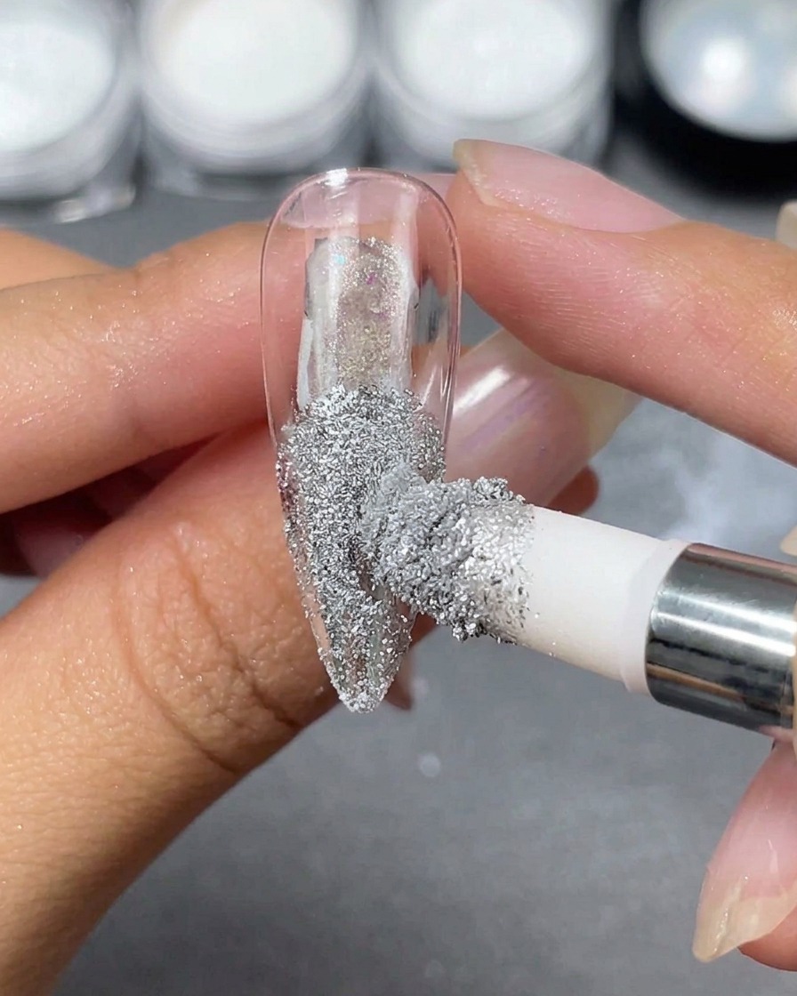 A sponge applicator depositing silver chrome powder onto a transparent stiletto nail tip held between fingers.
