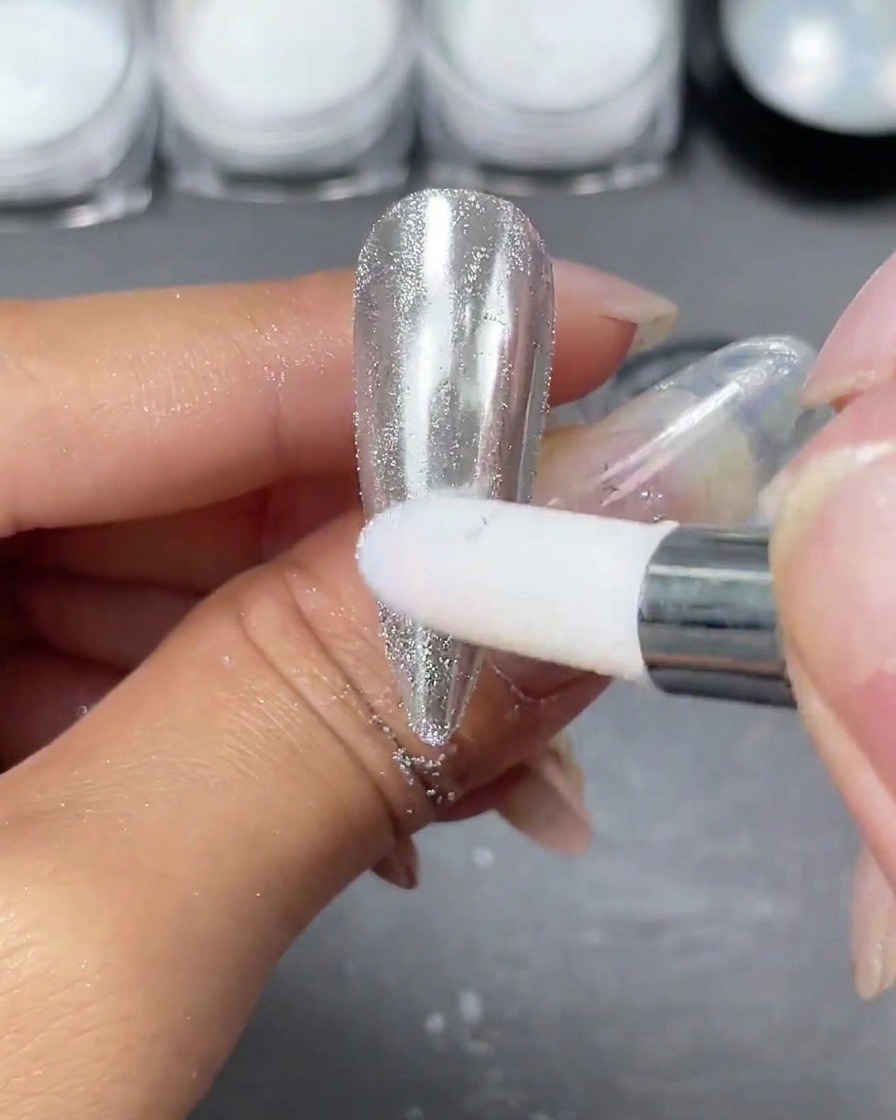 A sponge applicator rubbing silver chrome powder onto a clear nail tip until a bright, reflective mirror surface appears.