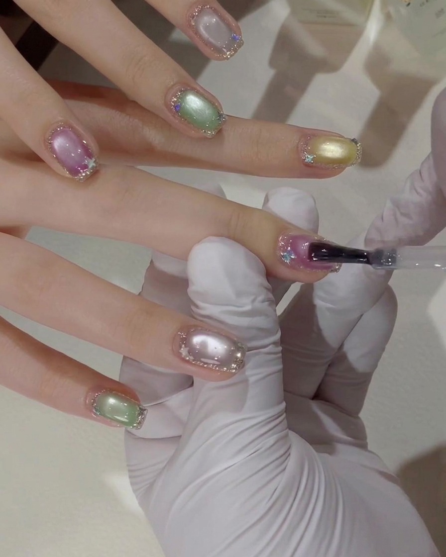 A close-up of a gloved hand using a gel brush to apply a clear top coat layer over a pink and purple cat-eye nail with silver glitter borders.