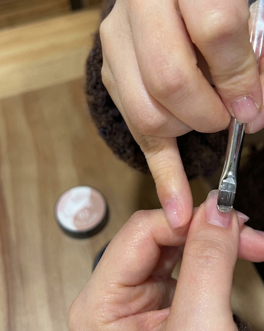 A close-up of a gel brush applying shimmering nude gold polish to a natural fingernail.
