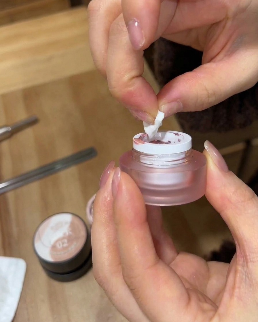 A close-up of hands using a torn piece of white makeup sponge to pick up pink gel polish from a small frosted jar.