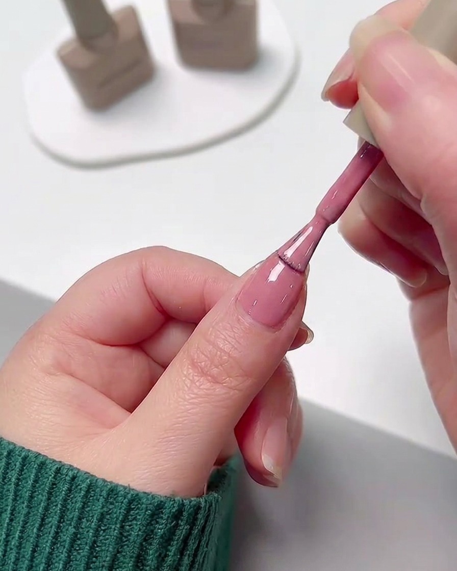 A hand applying a smooth layer of pink gel polish to a fingernail using the polish bottles brush.