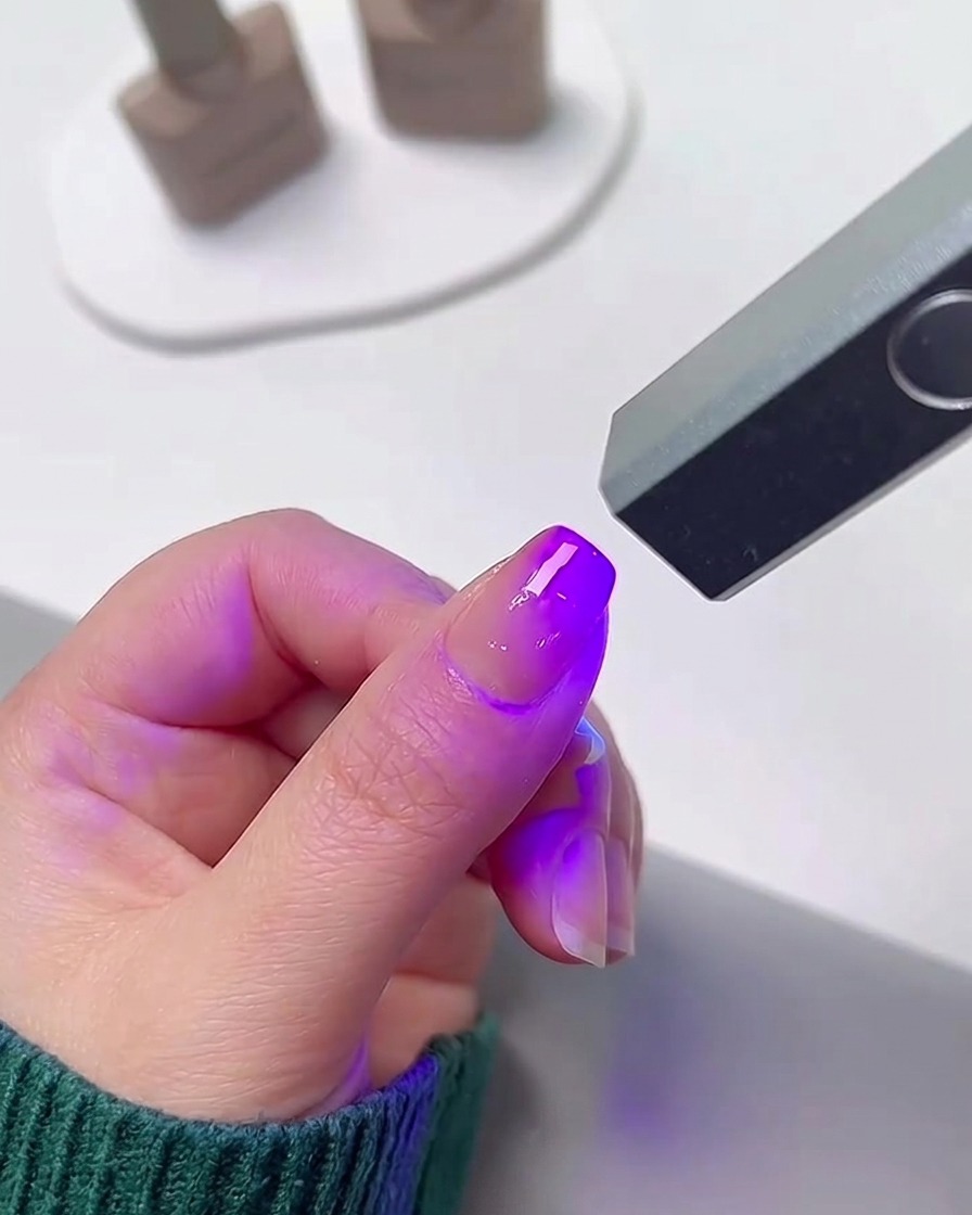 A finger positioned underneath a small, portable UV/LED nail lamp emitting a bright purple light to cure the gel polish.