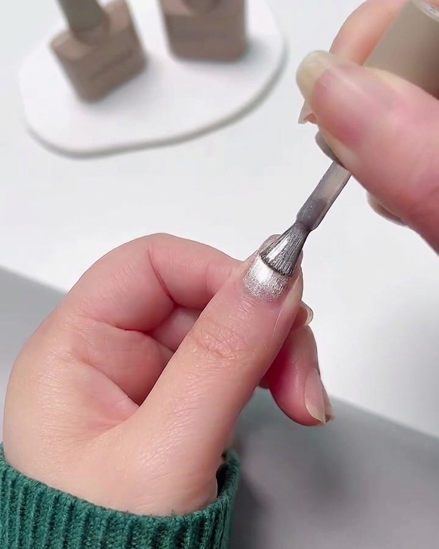 A hand applying a layer of shimmery silver cat eye gel polish over a cured pink gradient nail.