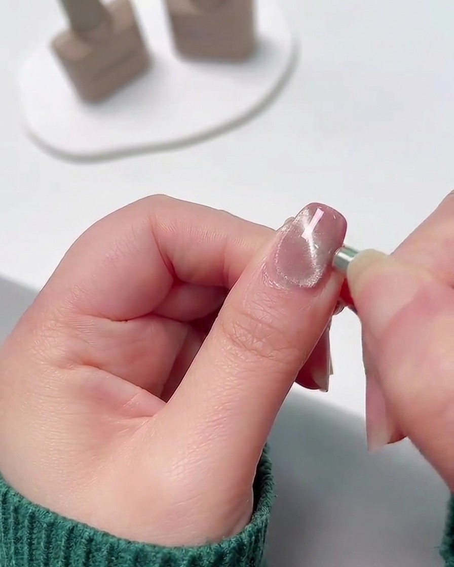 A close-up of a small cylindrical magnet being held against the side edge of a wet, shimmery gel nail to pull the metallic particles into a concentrated line.