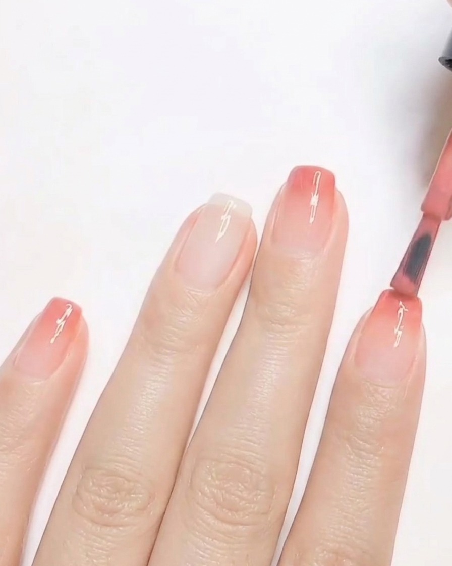 A nail brush applying a second coat of warm peach-toned gel polish to the tip of a fingernail to build an ombre gradient.