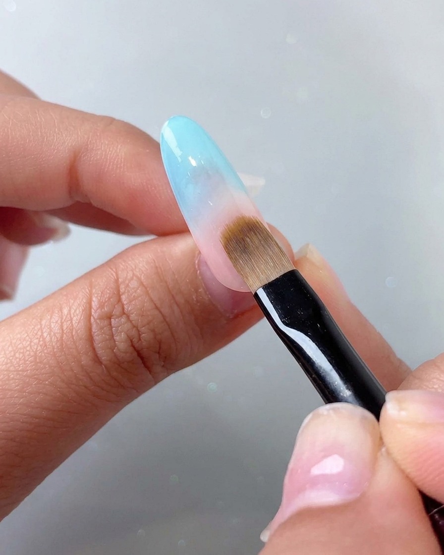 A patting brush blending a blue gradient effect onto the upper pinkish portion of the nail.