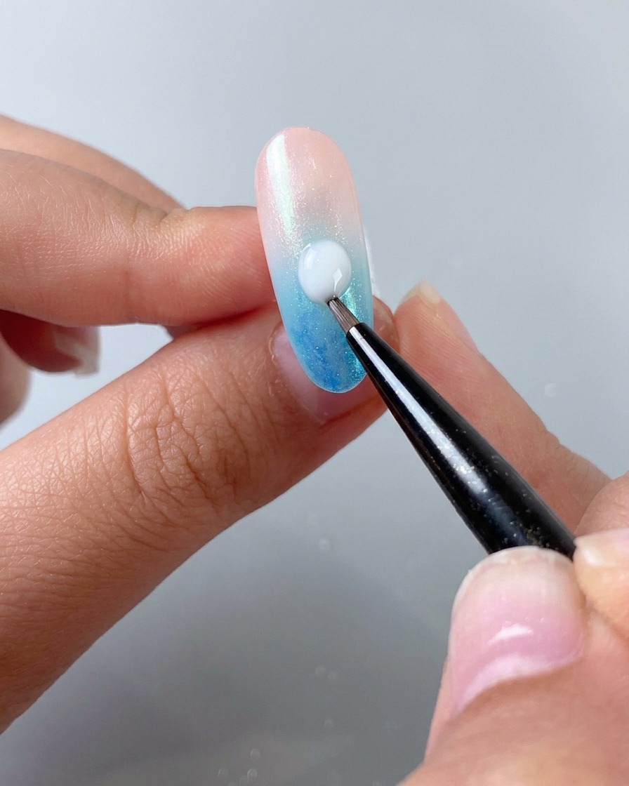 A fine nail art brush applying a drop of milky white self-leveling gel diagonally across a gradient nail.