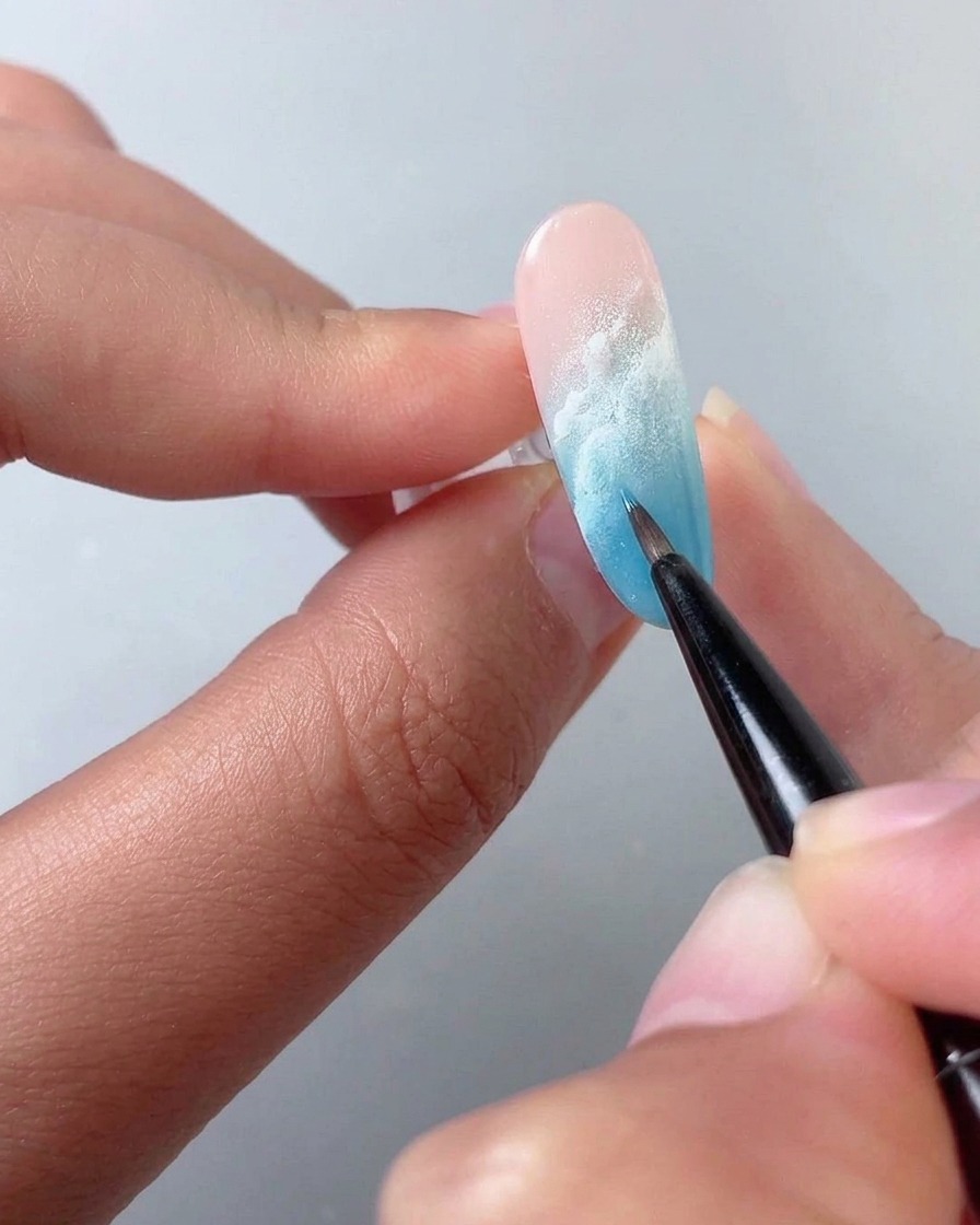 A fine brush blending white blooming liquid across a blue and pink gradient nail.
