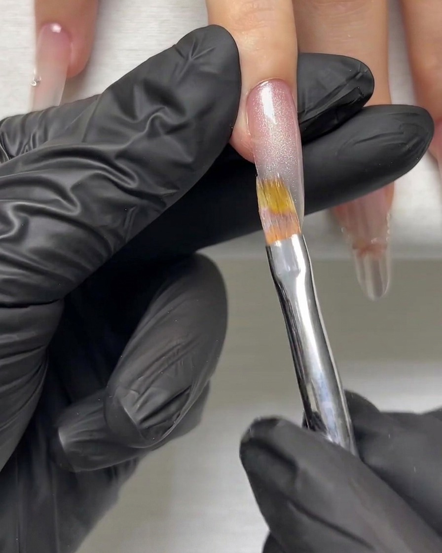 A gloved hand using a fine brush to smooth a silver shimmering gel layer on an elongated nail.