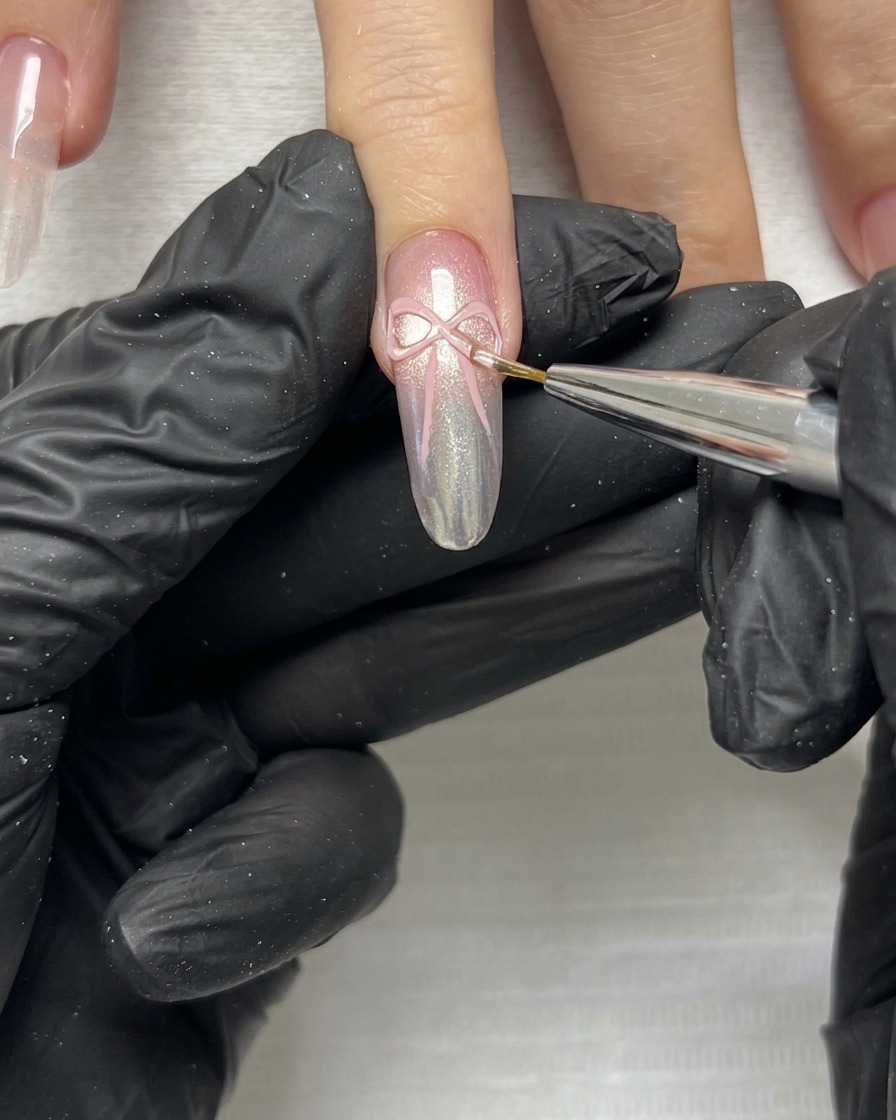 A close-up shot of a fine brush refining the delicate edges and tails of a pink 3D ribbon bow on a manicured nail.