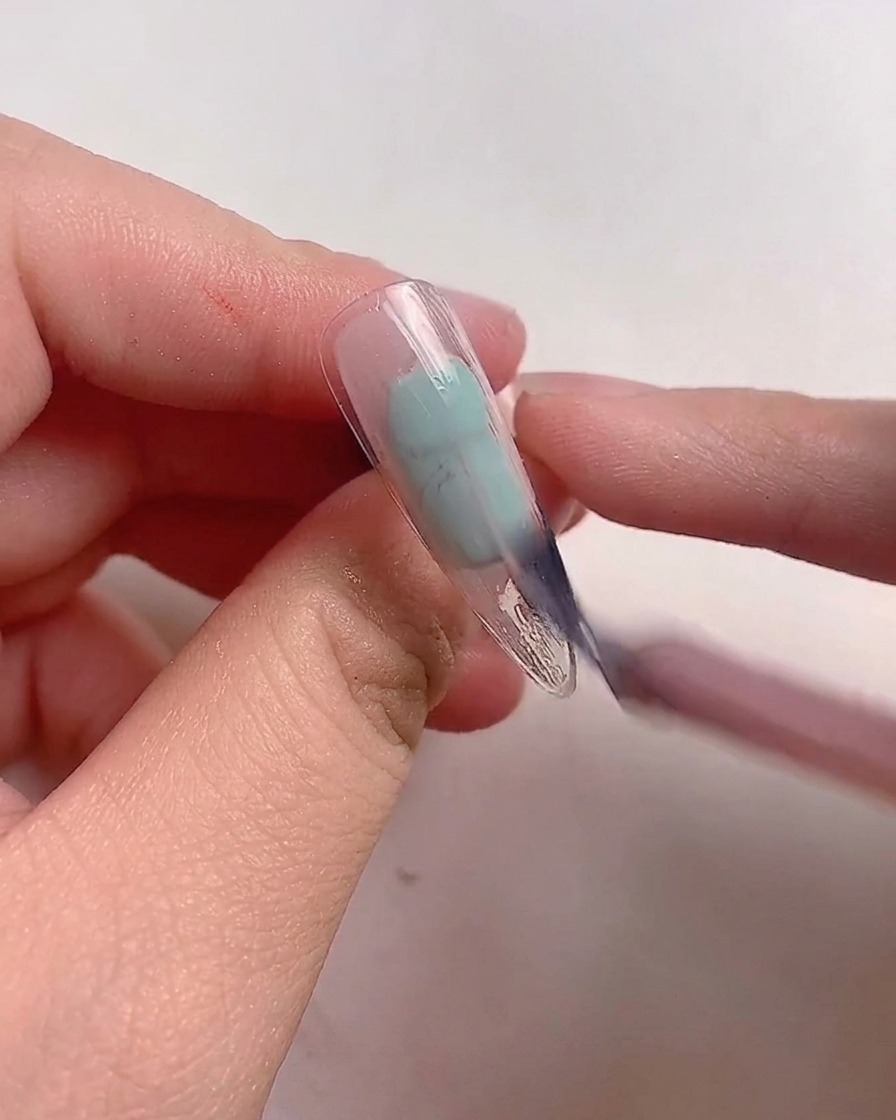 A brush applying mauve pink gel polish onto a clear nail tip.