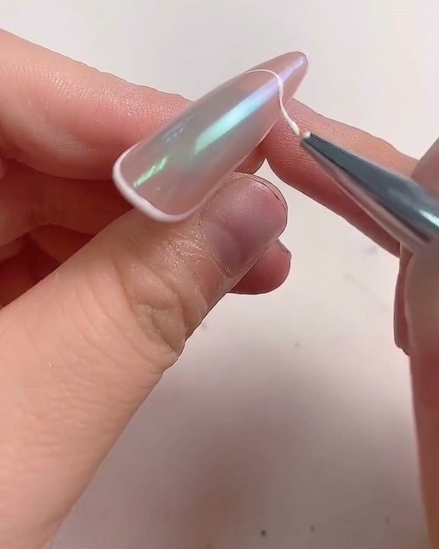 A fine liner brush applying a white gel line along the edge of an almond-shaped nail to create a French tip.