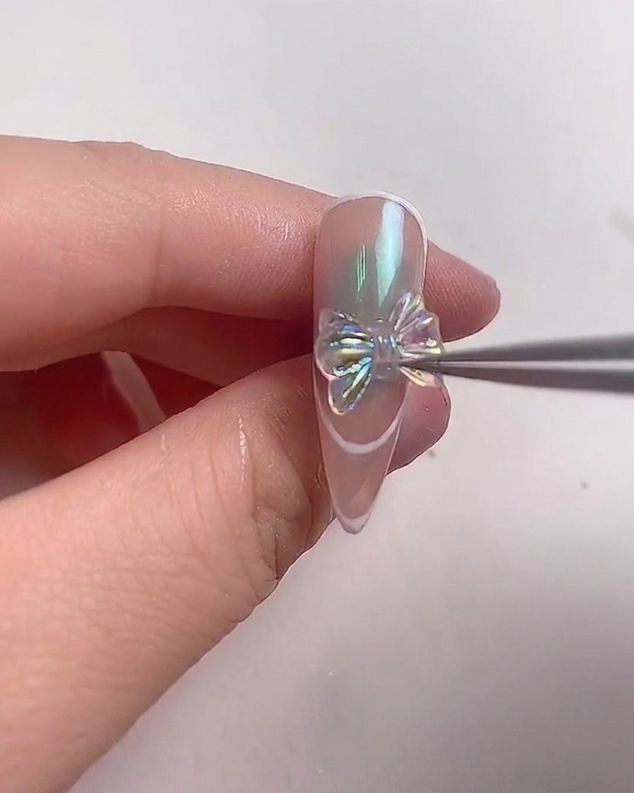 A pair of metal tweezers carefully positioning a sparkling iridescent crystal bow onto the center of an almond-shaped nail.
