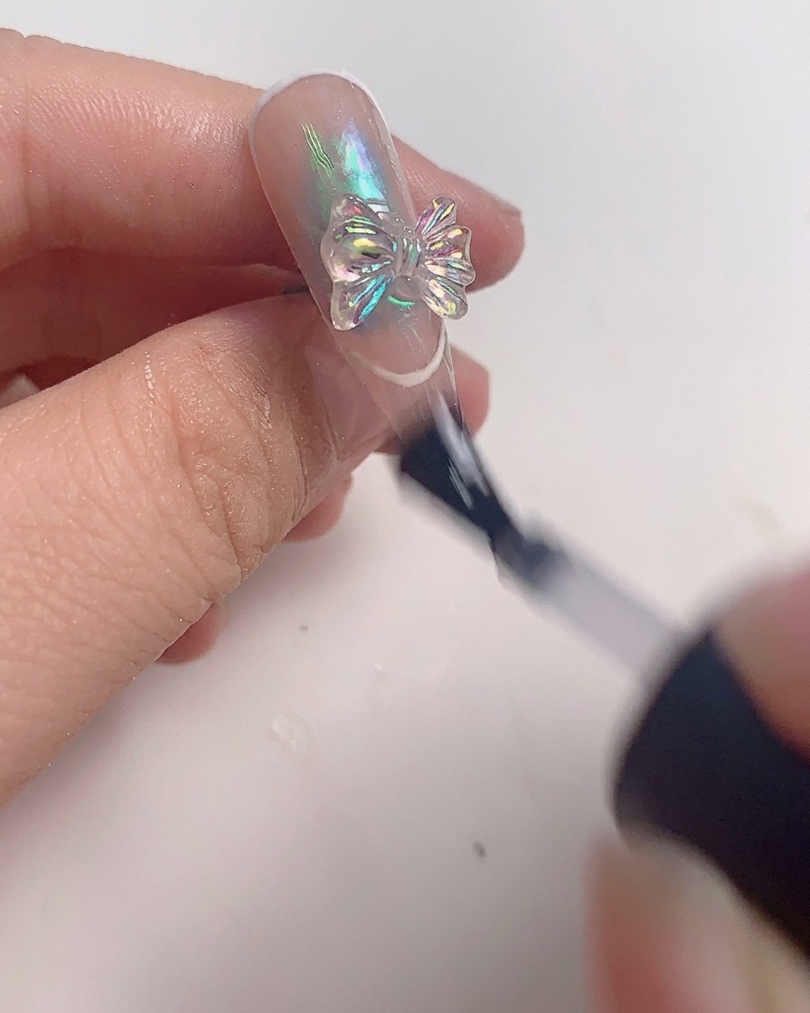 A brush applying a final layer of clear top coat around a crystal bow charm on a finished nail.