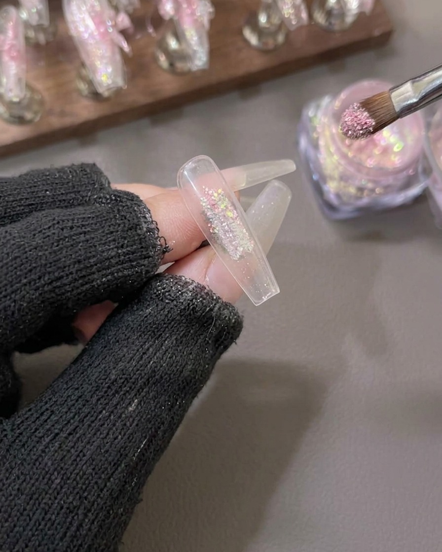 A brush applying sparkling pink and gold aurora flake powder to the center of a clear artificial nail tip.