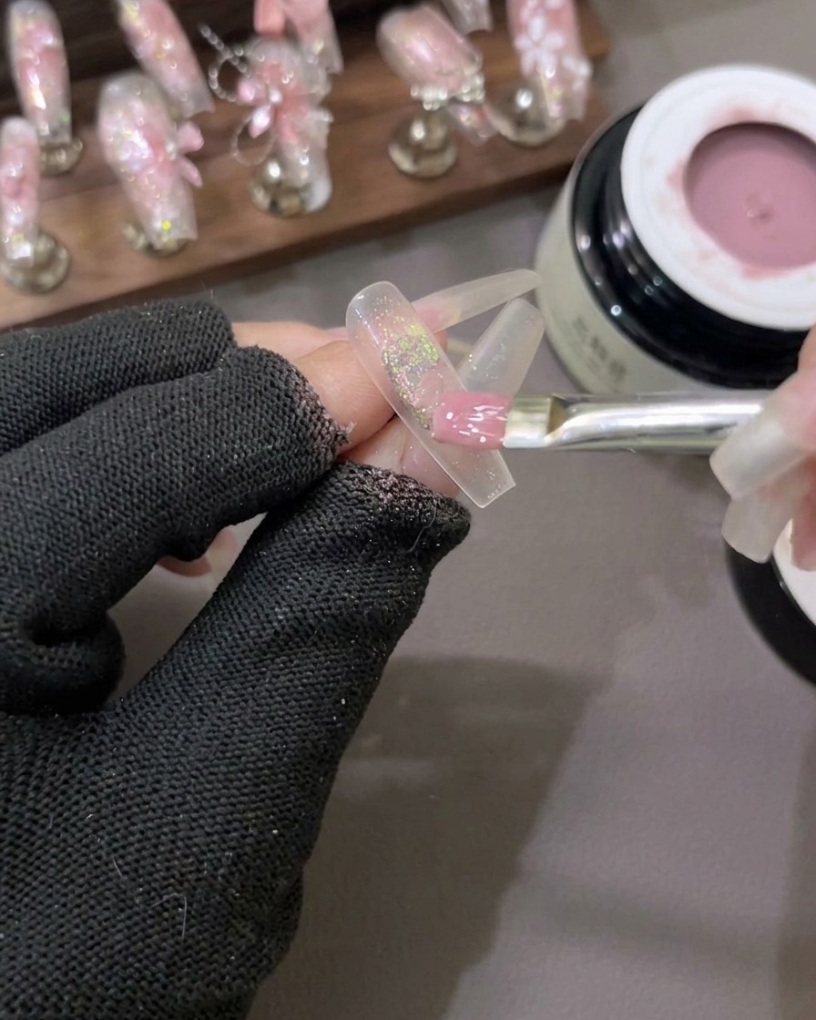 A flat gel brush spreading a semi-sheer milky pink gel polish over a nail with glitter flakes in the center.