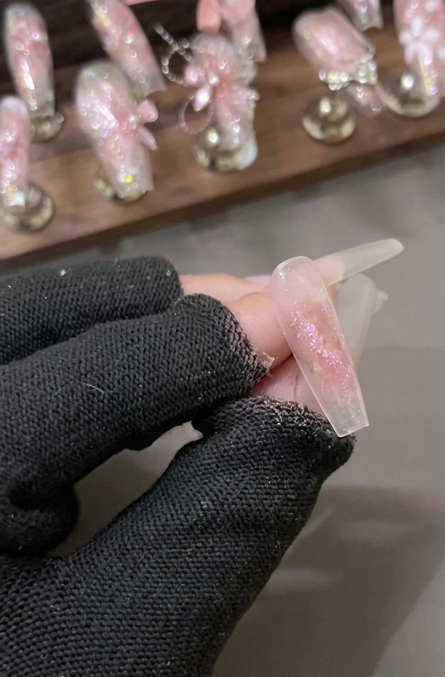A gloved hand holding a clear nail coated with a shimmering layer of pink shattered diamond glitter gel.