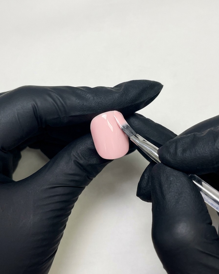 Gloved hands using a flat nail brush to apply skin pink gel polish to a prepped fingernail.
