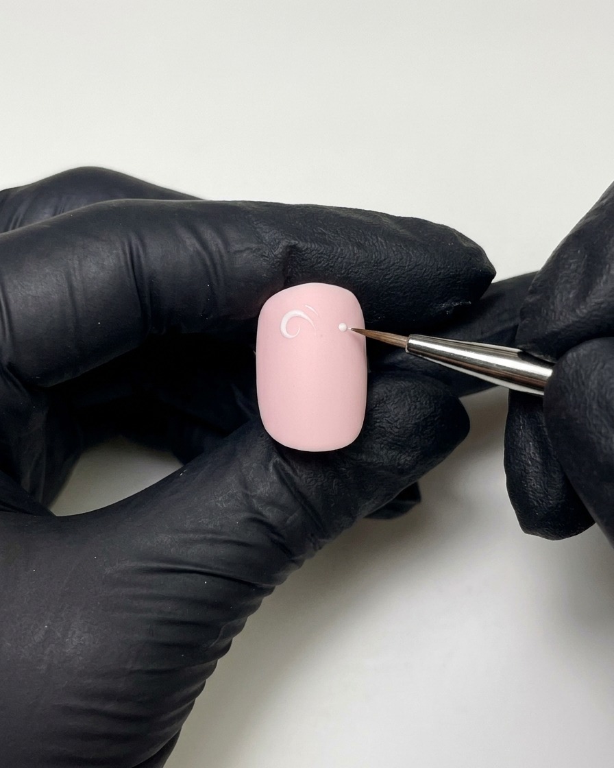 Gloved hands using a fine liner brush with builder gel to draw a white-pink loop and dot on a painted nail.