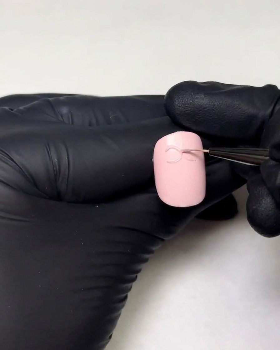 Gloved hands drawing the second loop of a 3D gel bow on a nail using a fine liner brush.