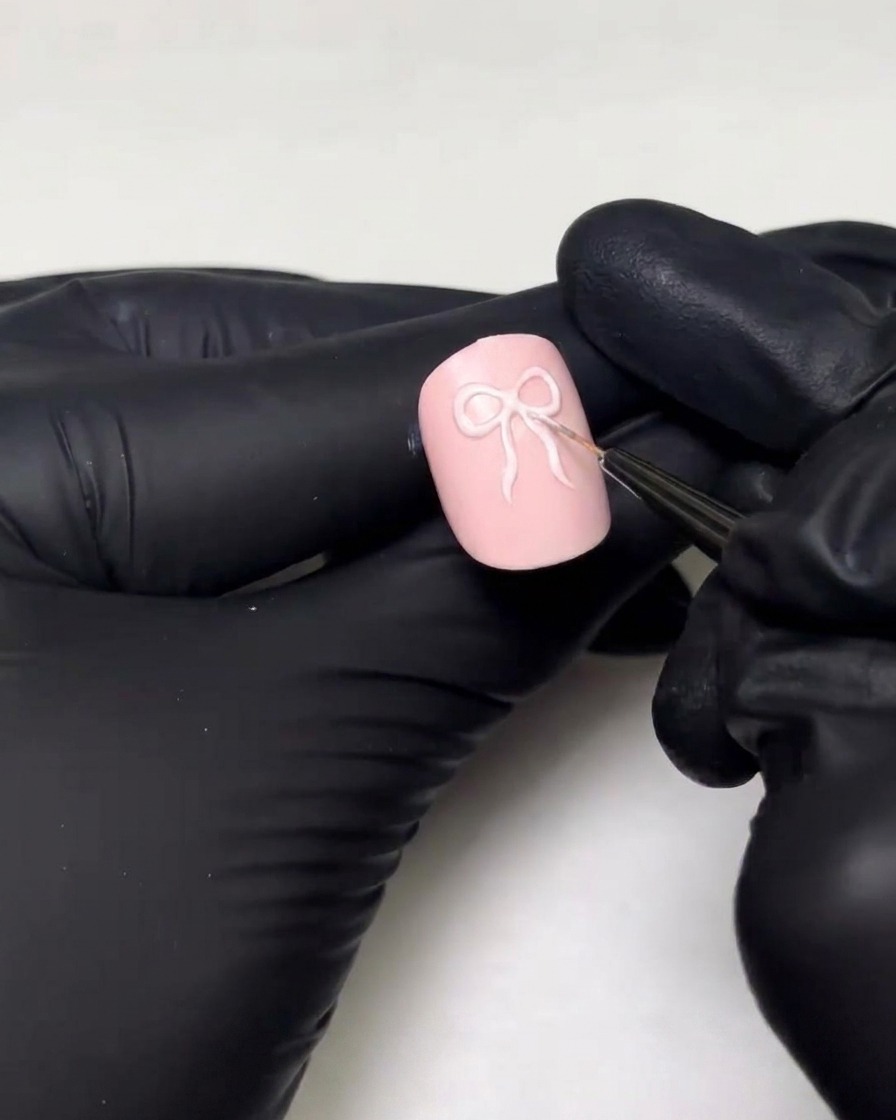 A gloved hand using a fine detailer brush to draw the loops and ribbon tails of a 3D bow design on a pink nail.