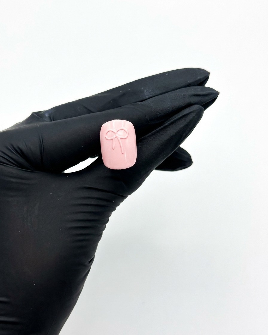 A gloved hand holding a pink press-on nail under a UV nail lamp to cure the 3D bow design.
