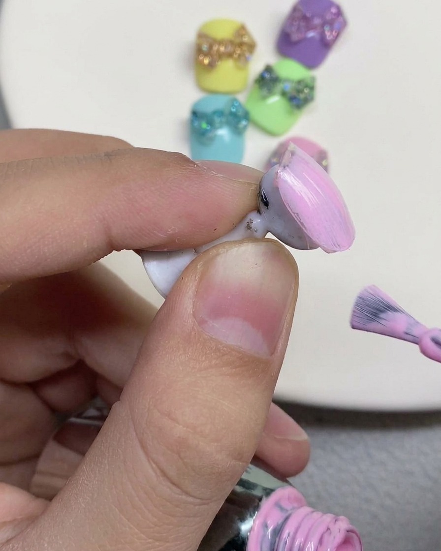 A nail brush painting a soft pastel pink gel polish onto the surface of a false nail displayed on a holder.